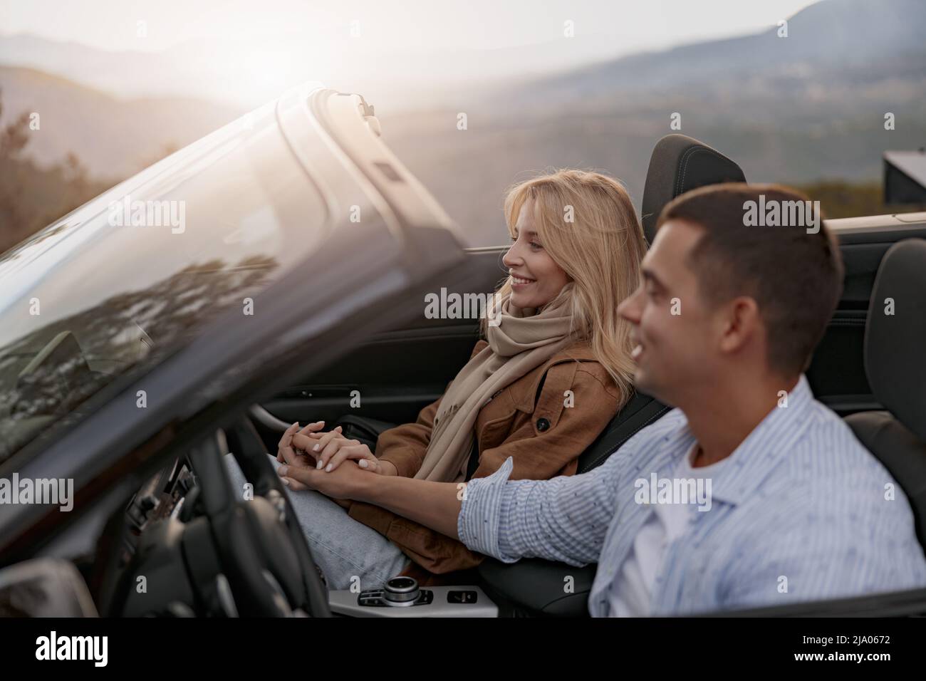 Cheerful pretty blond female smiling and holding hands with boyfriend in a convertible car Stock Photo