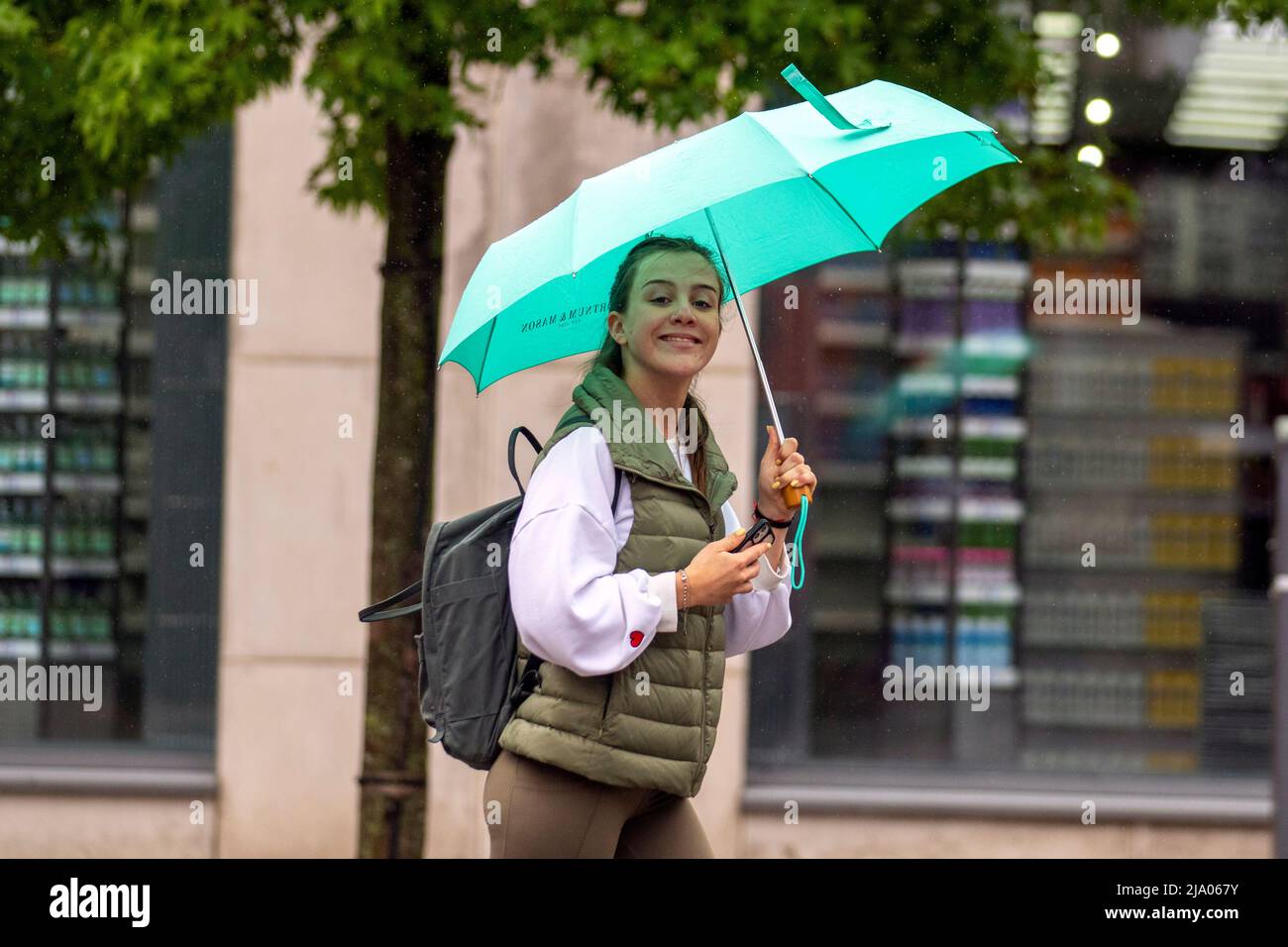 Smiling young woman with green umbrella in the rainPreston, Lancashire.  UK Weather May 2022.  No signs of summer as yet another rainy start to the day for shops, shoppers and shopping in Preston. A dull and wet start to today, with spells of rain moving in.  Credit. MediaWorldImages/AlamyLiveNews Stock Photo