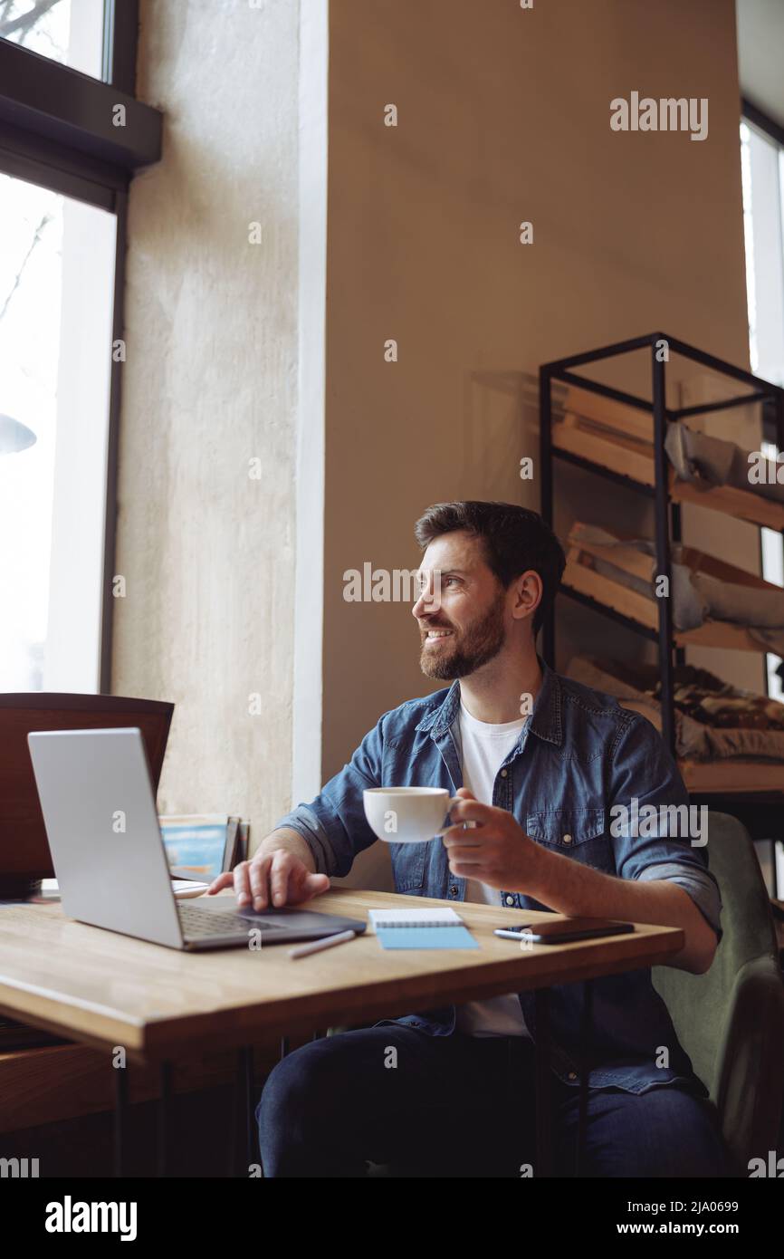 Attractive Caucasian male freelancer sitting in cafe, looking at window, having coffee at laptop. Stock Photo