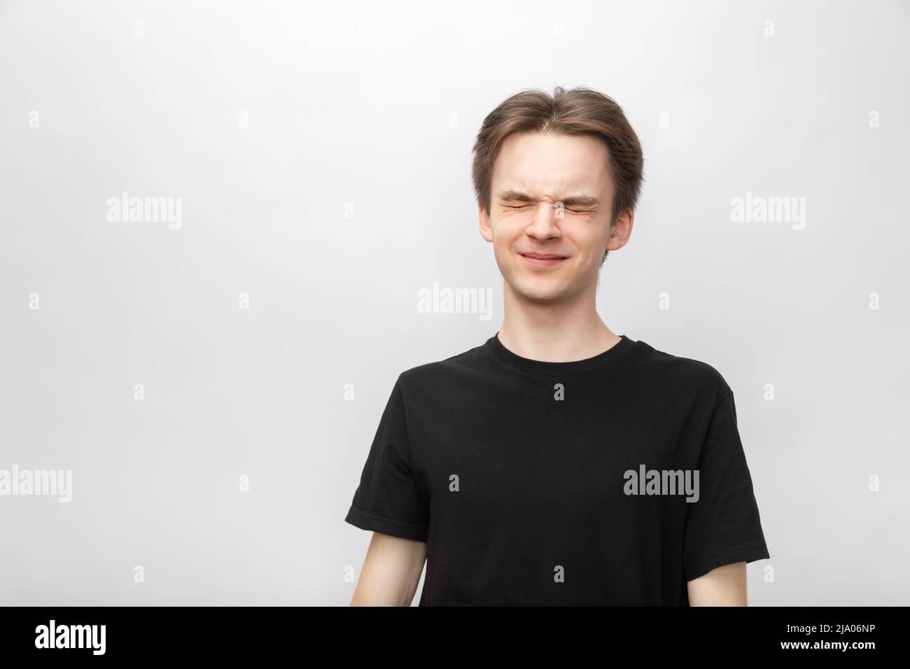 Portrait of annoyed young man wearing black tshirt disturbed from loud awful sound or noise tightly narrowing his eyes. Studio shot on gray background Stock Photo