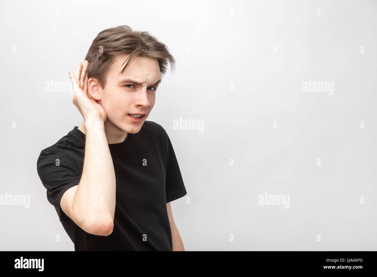 Portrait of young man wearing black tshirt listening holding hand near ear looking at camera. Studio shot on gray background Stock Photo