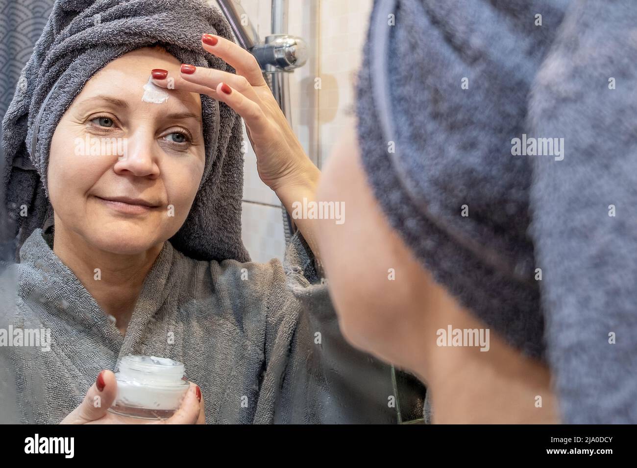 A young smiling woman in a towel applies face cream. The concept of skin rejuvenation. The concept of skin care Stock Photo