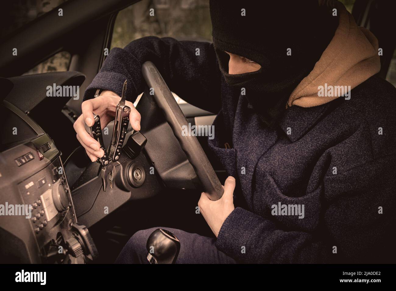 A car thief is trying to open the ignition lock with a tool. The car thief hid his face in a balaclava Stock Photo