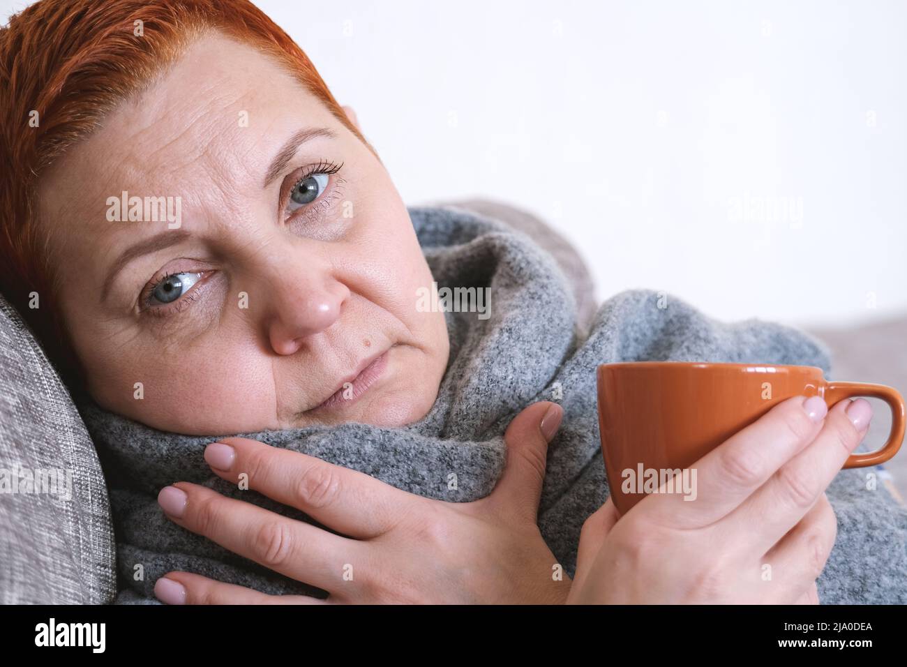 A young woman with a cold or flu-like illness. Portrait of an unhealthy girl with a scarf around her neck to warm a sore throat, drinking tea. Colds a Stock Photo