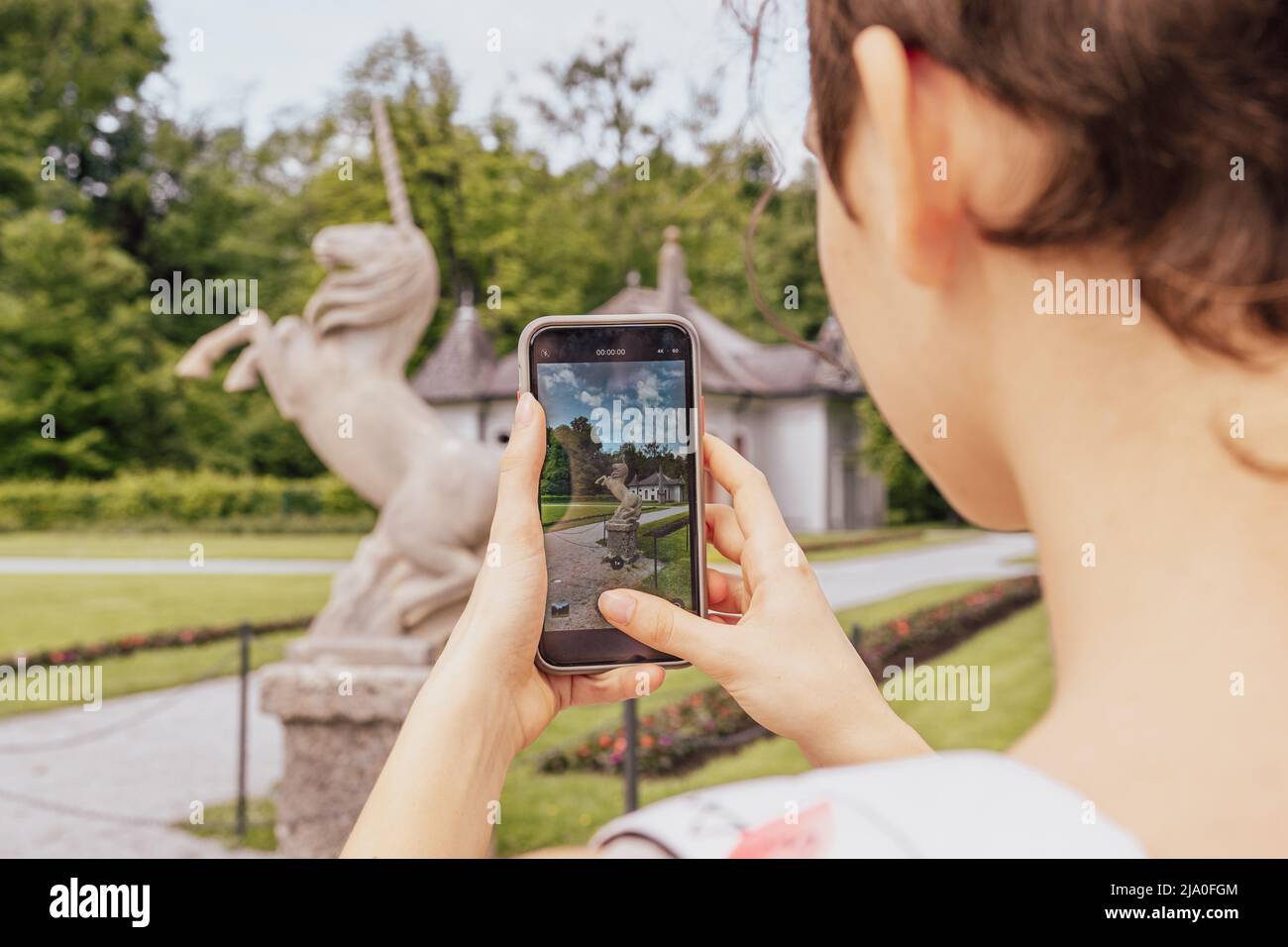 unrecognizable girl from back takes pictures on smartphone of historical landmarks, stone sculpture in shape of unicorn, in Hellbrunn park. Austria, Stock Photo