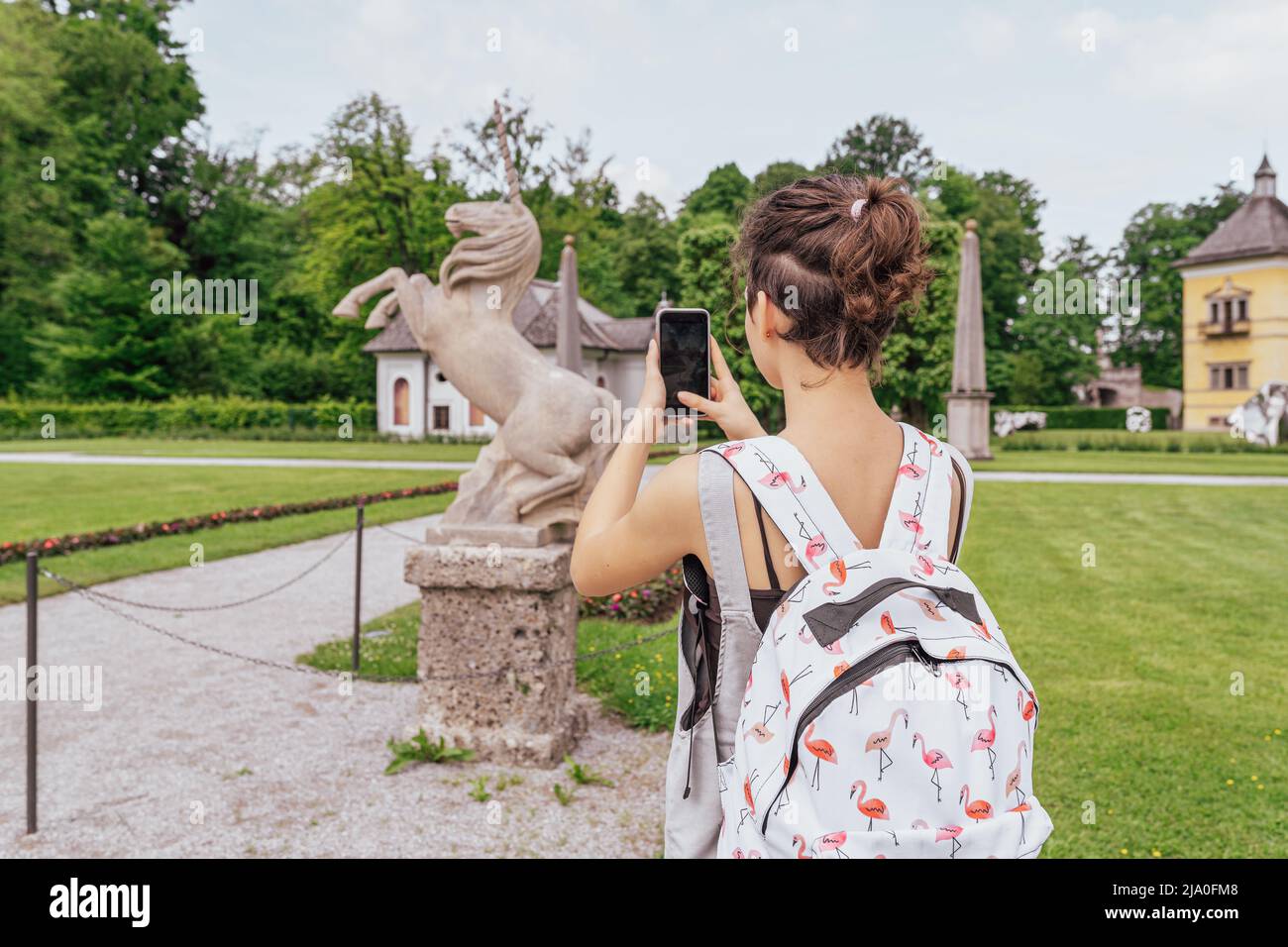 unrecognizable girl from back takes pictures on smartphone of historical landmarks, stone sculpture in shape of unicorn, in Hellbrunn park. Austria, Stock Photo