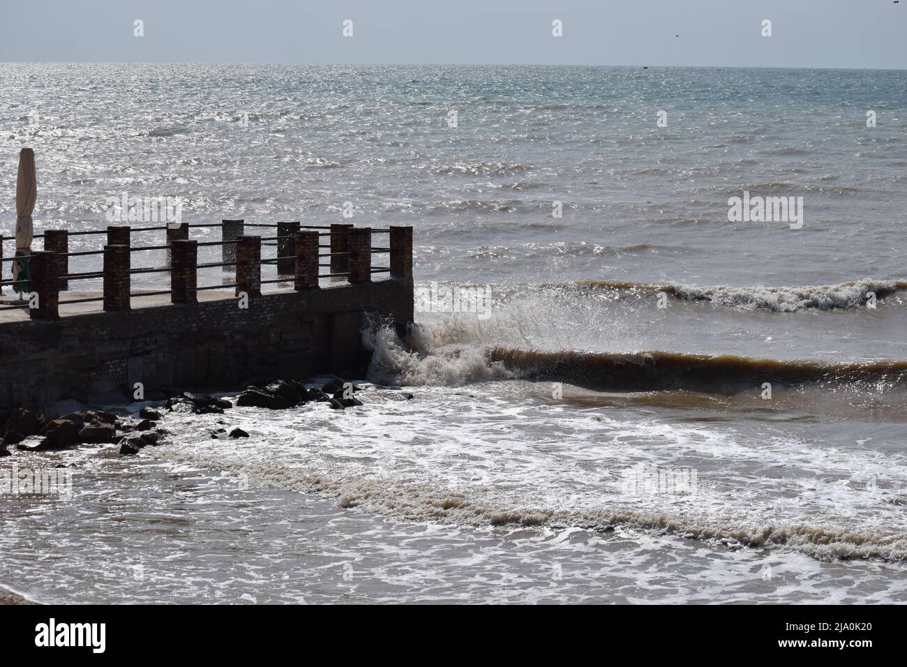 big sea waves splashing against a pier. The pier cuts the sea waves off the coast during a stormy sunny day. Stock Photo