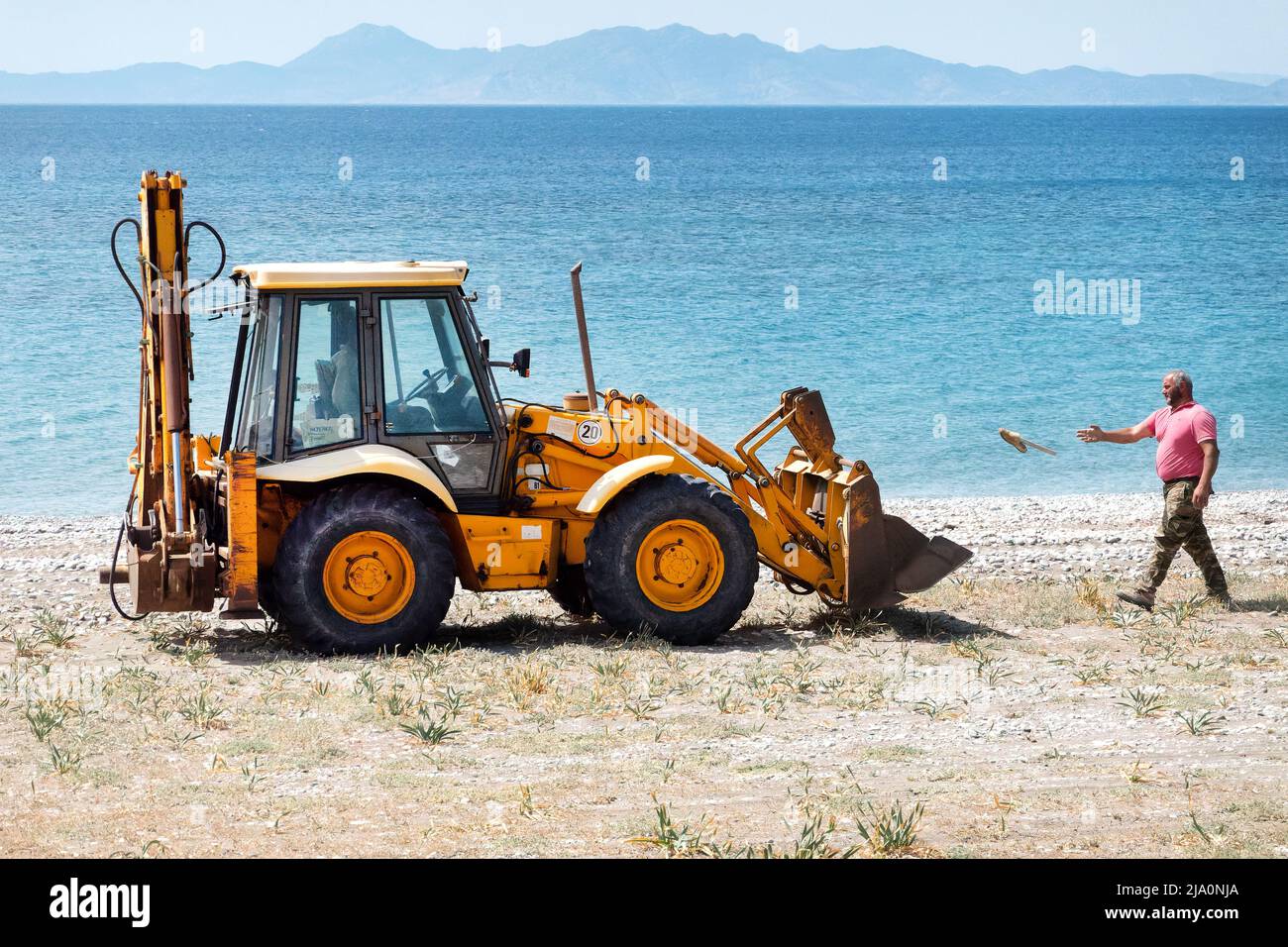 A workman throws litter into a JCB diggers skip whilst clearing and cleaning up discarded rubbish on a beach near Rhodes City, Rhodes Greece. Stock Photo