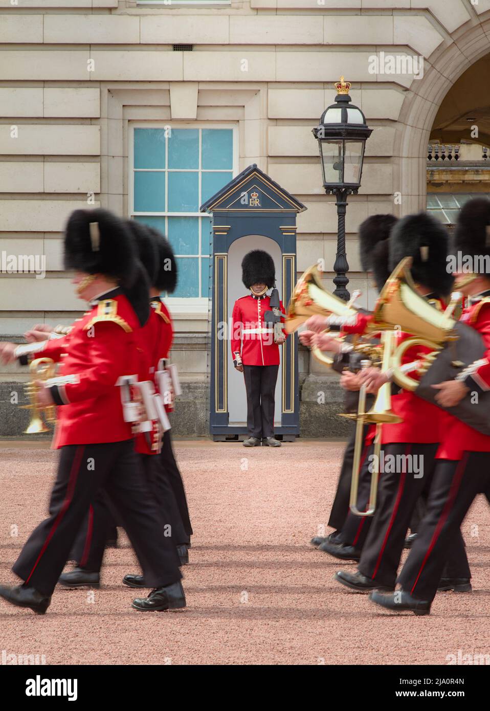 Regimental Band Or Corps Of Drums Of The Queens Guard Marching Past A New Guard Positioned In His Sentry Box Standing To Attention, Buckingham Palace Stock Photo