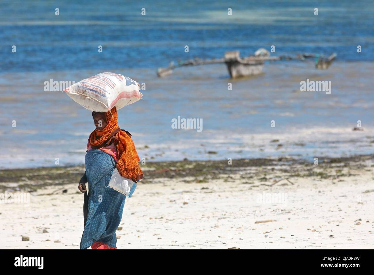 A woman carrying a sack of seaweed on her head at the Jambiani beach with a traditional 'Ngalawa' boat in the background, Zanzibar, Tanzania, Africa. Stock Photo