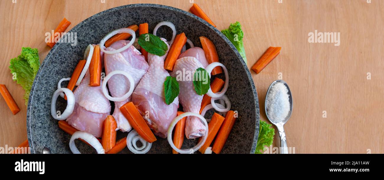 Raw chicken drumsticks with carrots, onions and green herbs in frying pan top view. Fresh meat