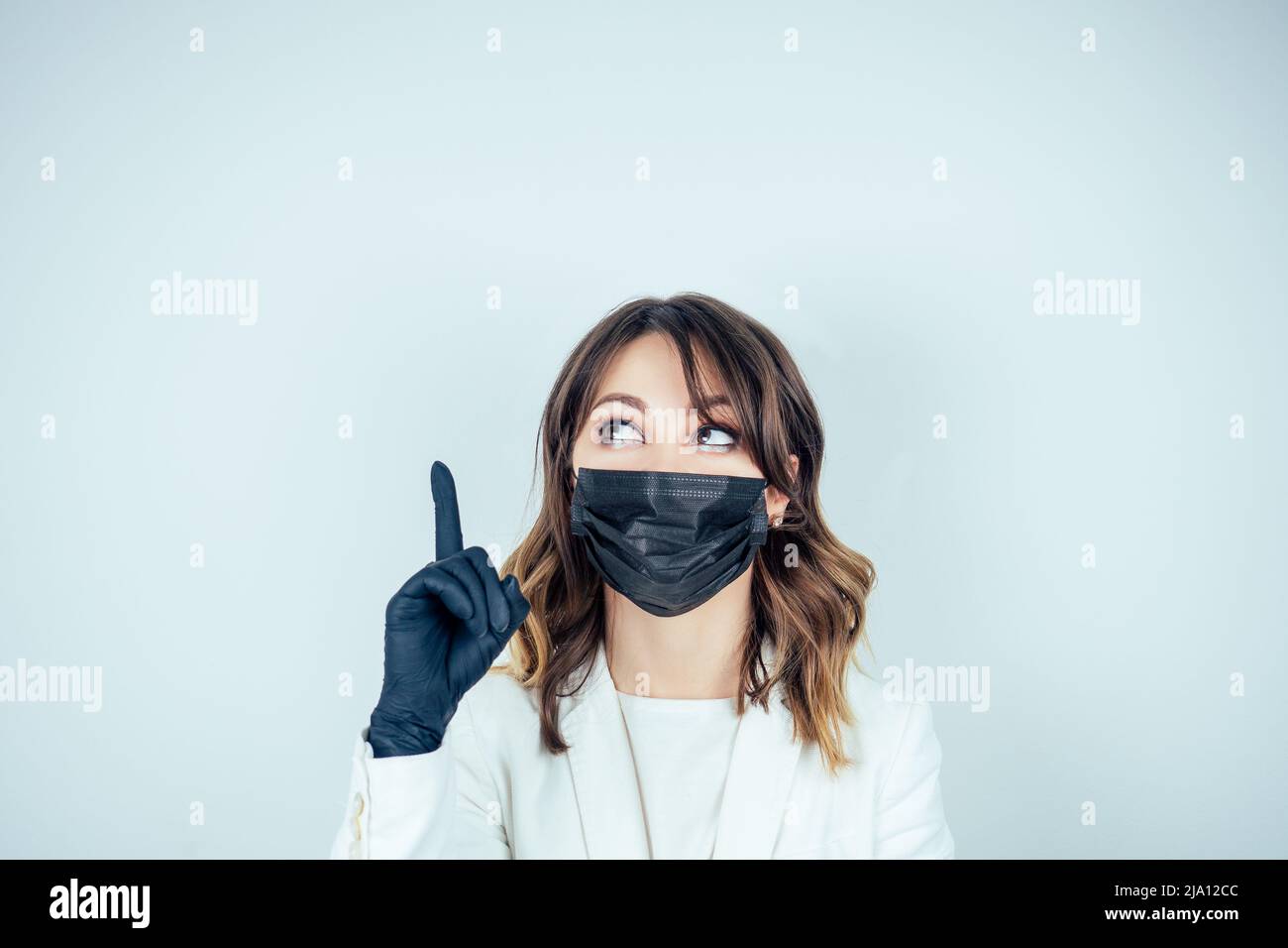 portrait of a beautiful and young doctor woman in white medical gown , black rubber gloves and black mask on face finger pointing up on a white Stock Photo