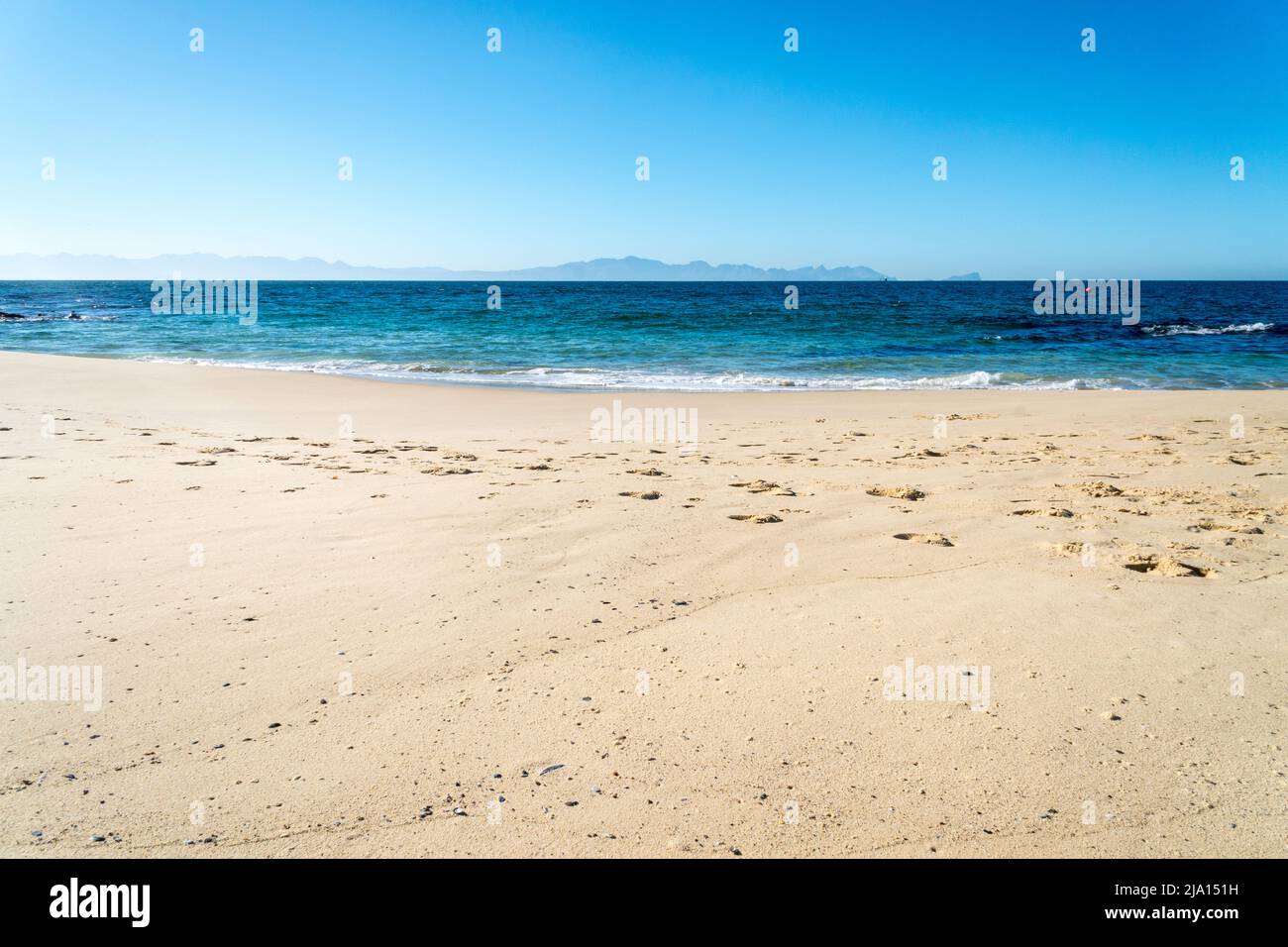 empty white sand beach with no people and calm sea with cloudless blue sky and mountains in the background concept weather and climate Stock Photo