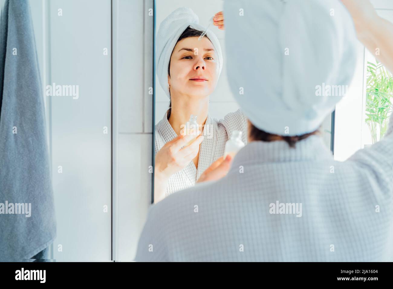 Mixed raced woman in bath spa towel applying rejuvenation moisturizing serum on her face looking at the mirror at home after hot bath and shower. Beau Stock Photo