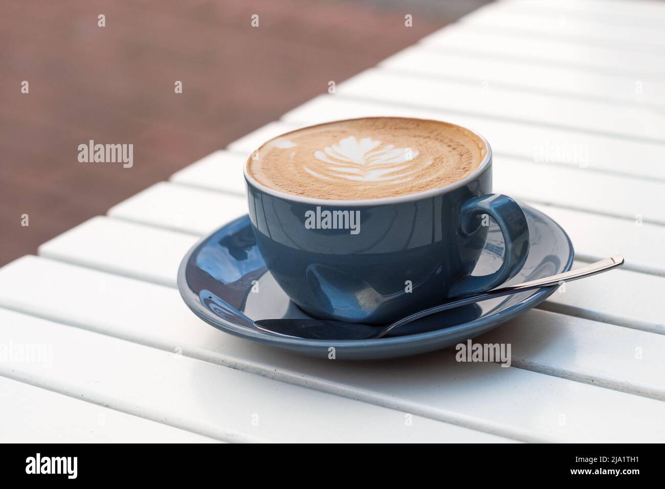 Shallow focus on the froth of flat white coffee in a blue cup and saucer with a teaspoon on the saucer. The coffee is on a white wooden slated table o Stock Photo