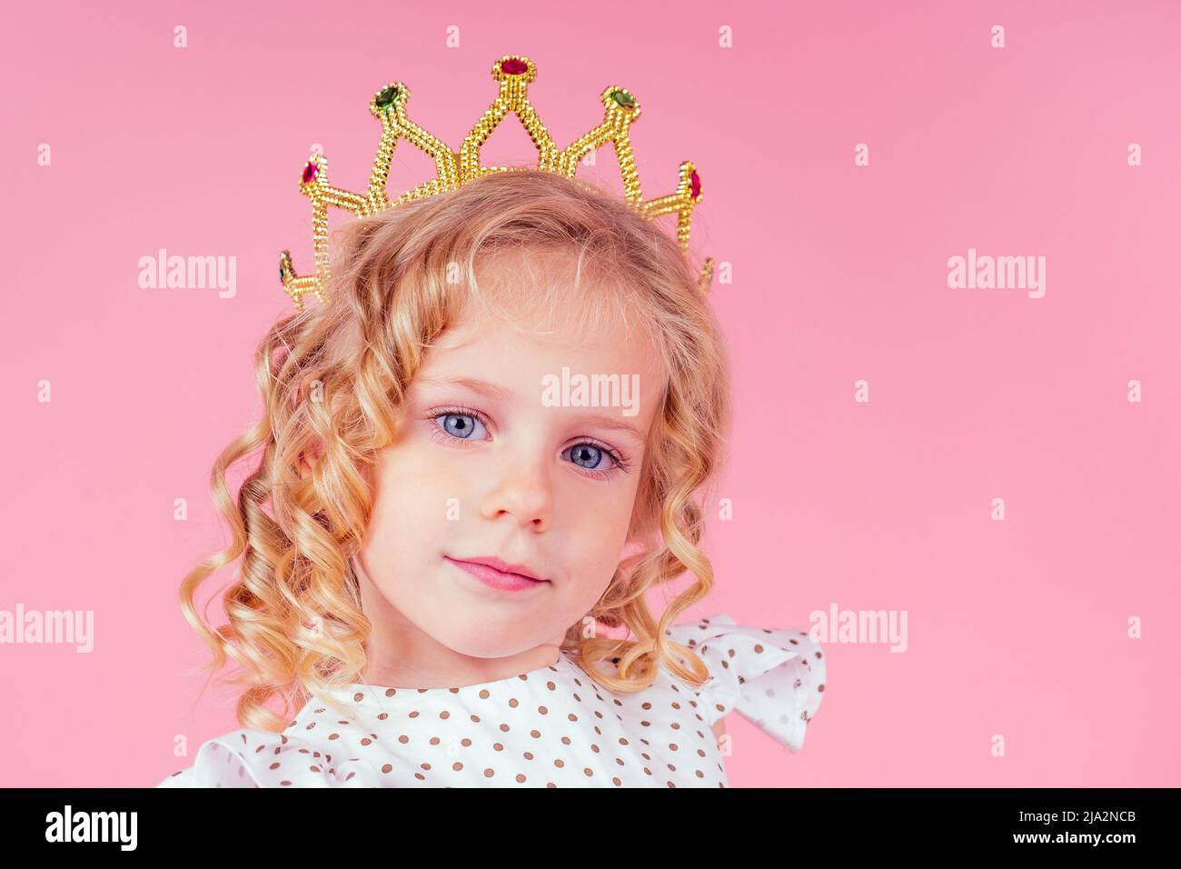 little girl beauty queen blue eyes, curls blonde hairstyle with a tiara crown on her head in a cute white dress in peas posing in the studio on a pink Stock Photo