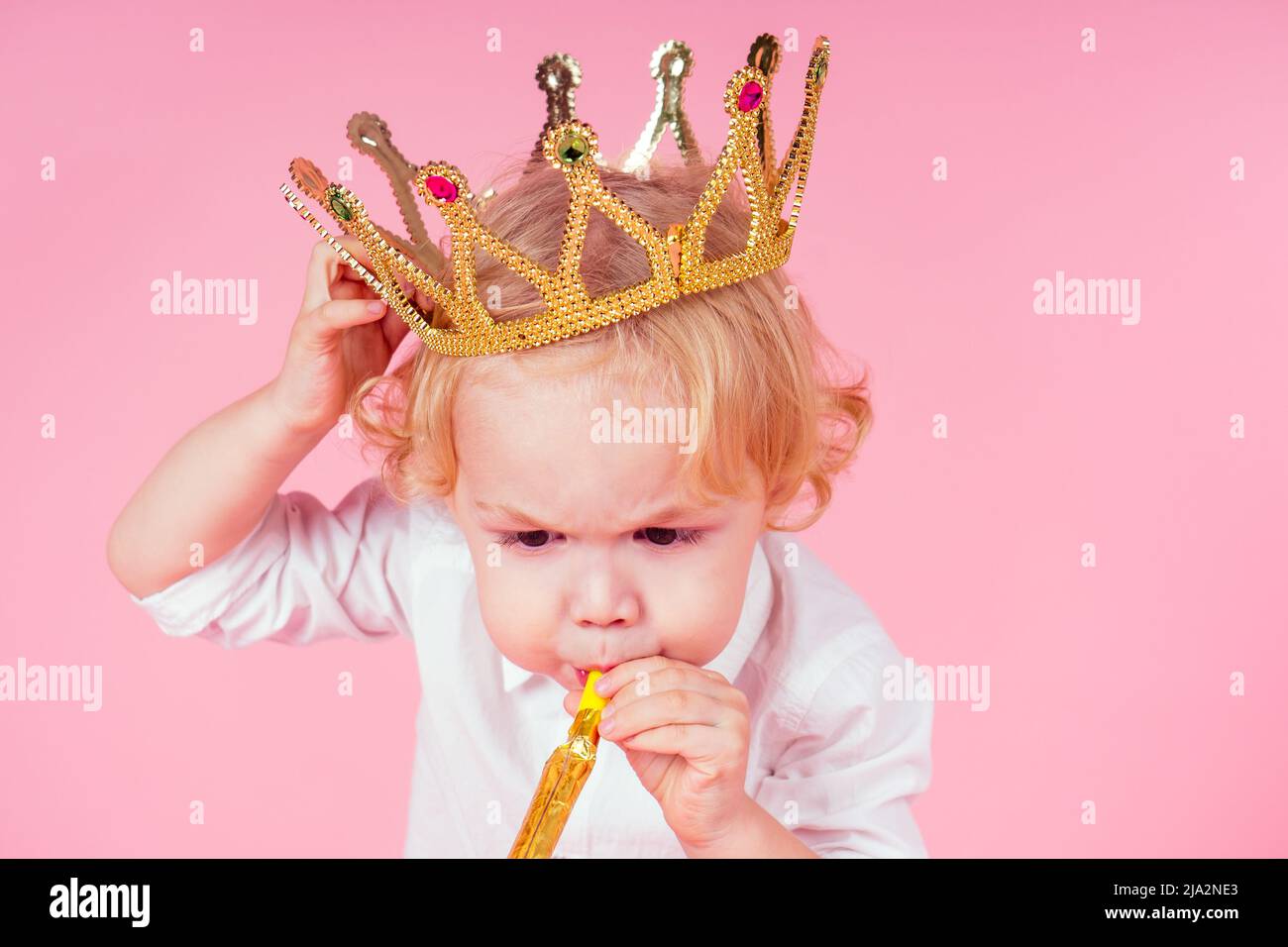 little blond boy with golden crown head curls hairstyle 4-5 year old in studio on pink background blowing noisemakers horn-whistle a birthday party Stock Photo