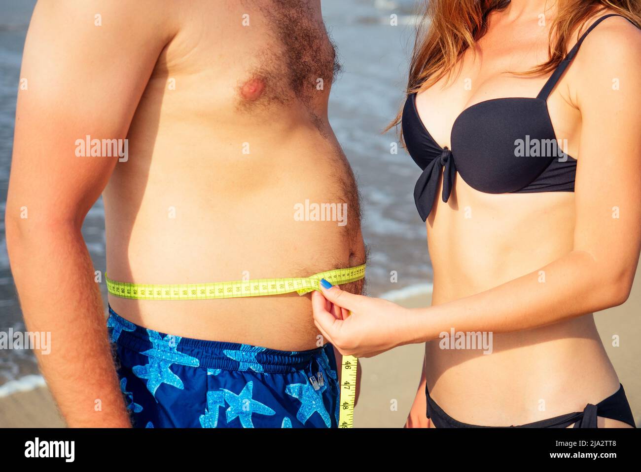 slender woman measures the volumes of a fat man on the beach Stock Photo