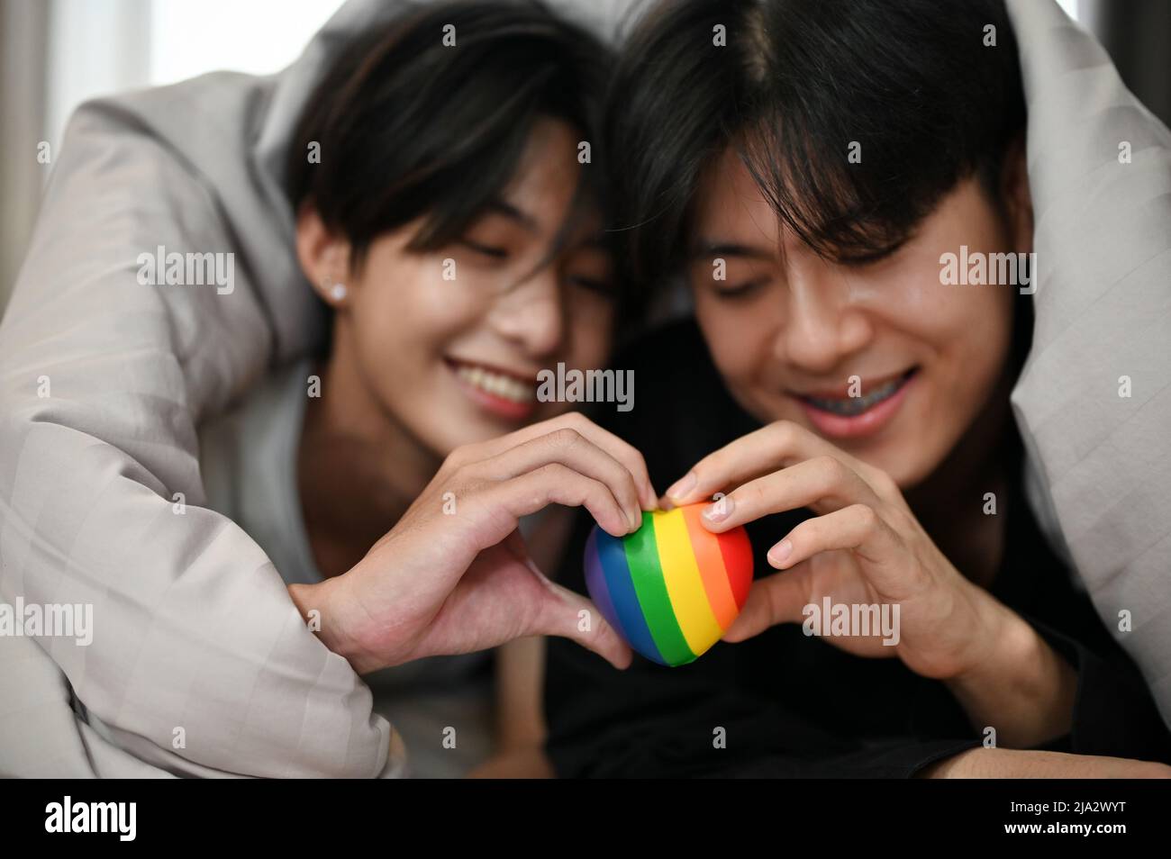 Lovely asian young gay couples lying on bed under the blanket and holding a rainbow LGBT heart symbol together. Happy gay couples, LGBT gay men. Stock Photo