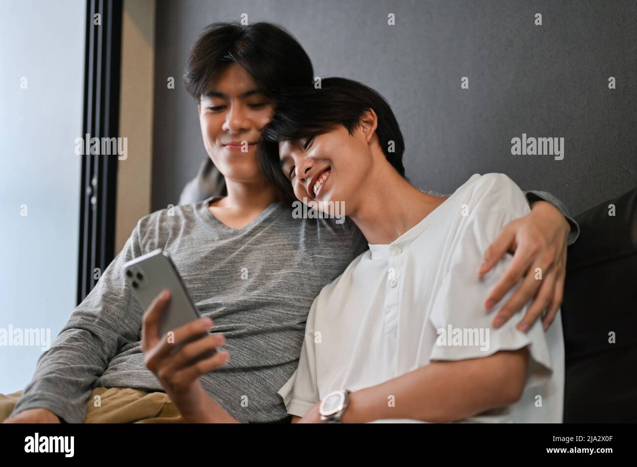 A gay man arm around his boyfriend shoulders, relaxes in living room, watching something on smartphone together. LGBT couples concept Stock Photo