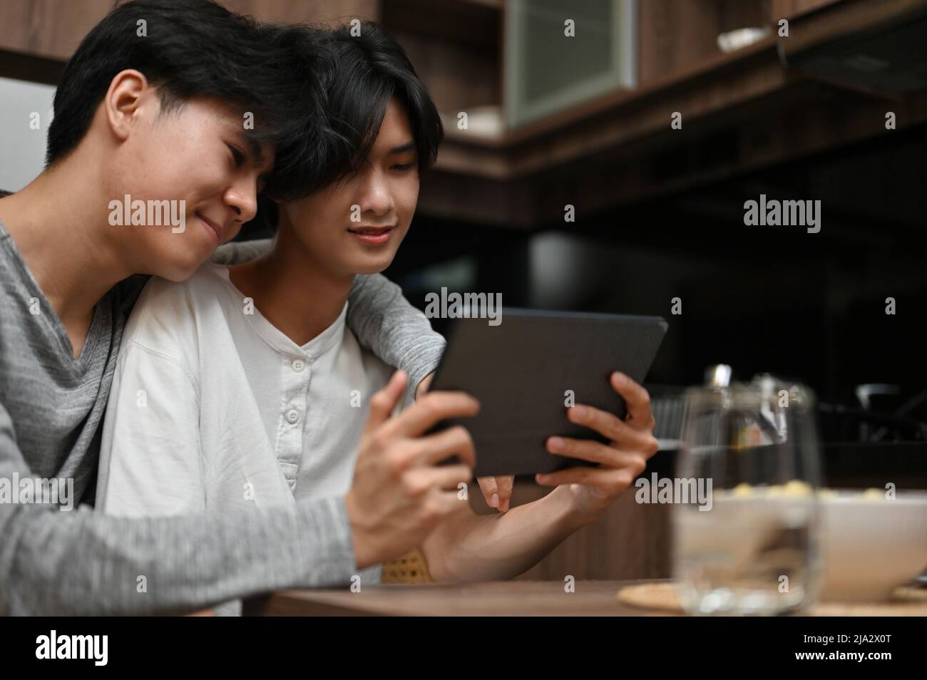 Happy asian young gay men couple showing some LGBT love affection, watching movie on tablet together in the kitchen. Stock Photo