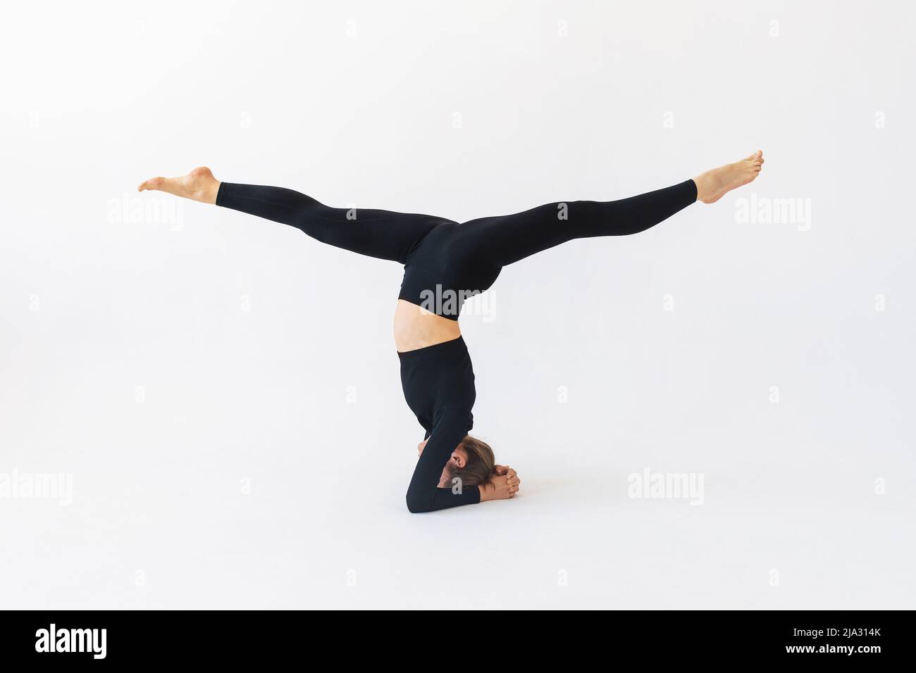 Woman in black sportswear practicing yoga doing shirshasana exercises with hanumanasana, headstand Stock Photo