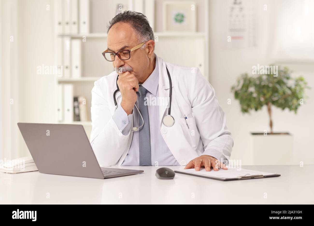 Mature male doctor sitting in an office and looking at a laptop computer Stock Photo