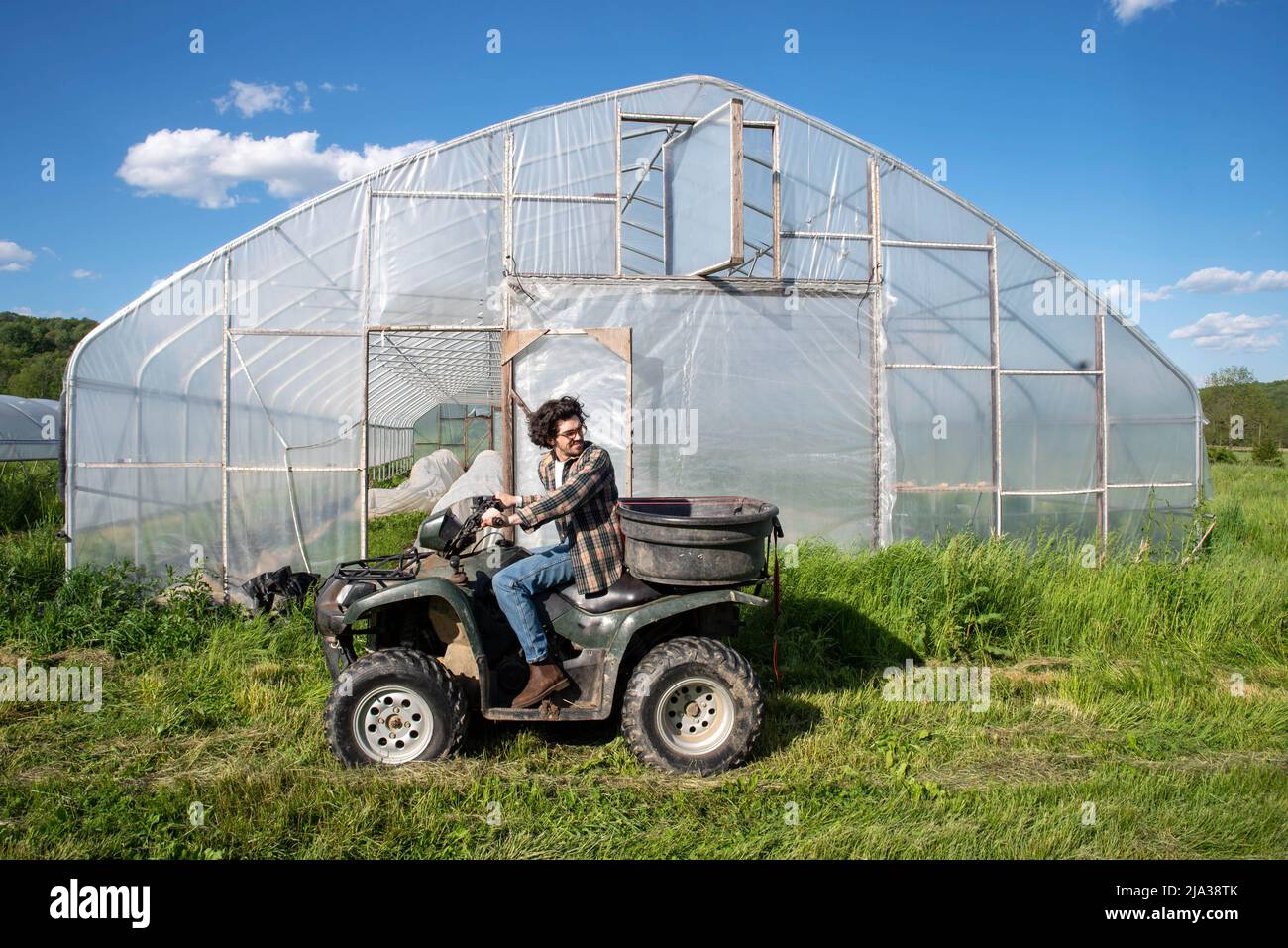 Young caucasian male farmer drives an atv by a vegetable garden greenhouse on an organic farm. Greenhouse door is open with covered rows of vegetables Stock Photo