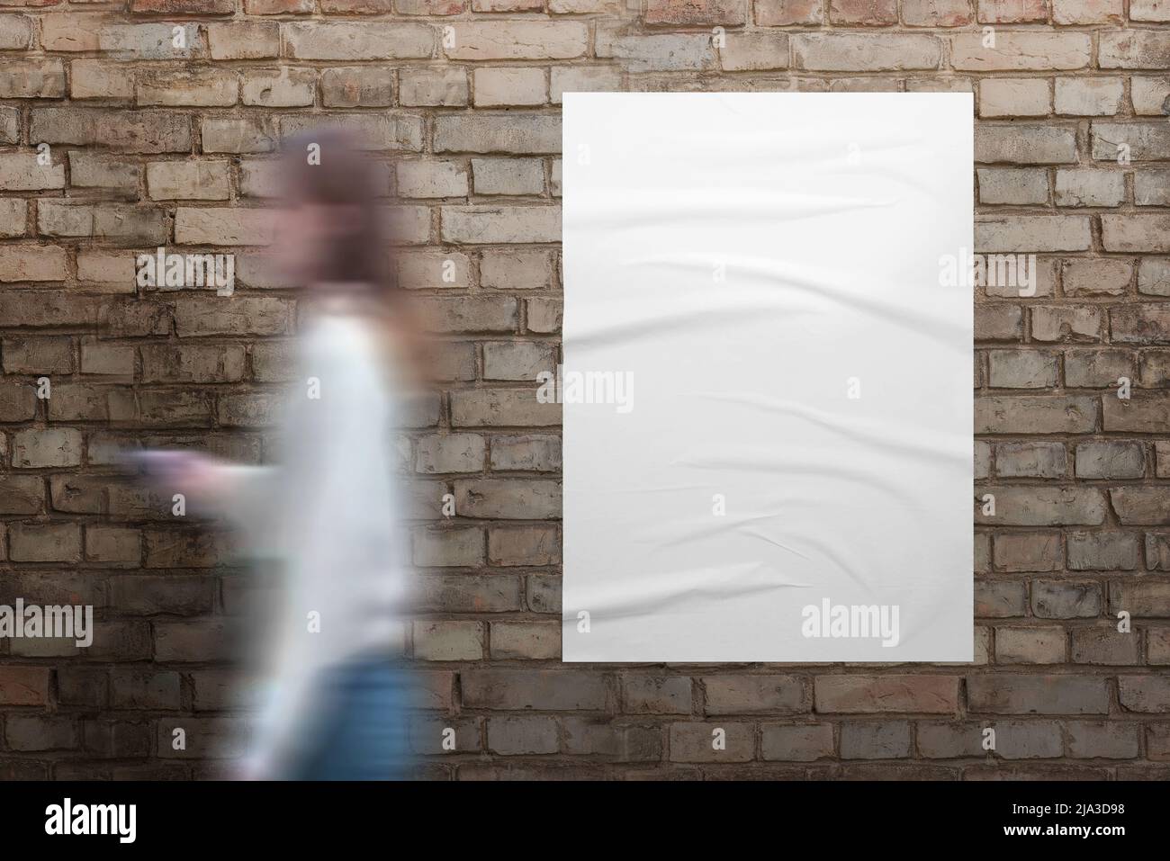 Girl walks past a white poster on brick street wall. Blank white poster with crumpled texture for design presentation Stock Photo