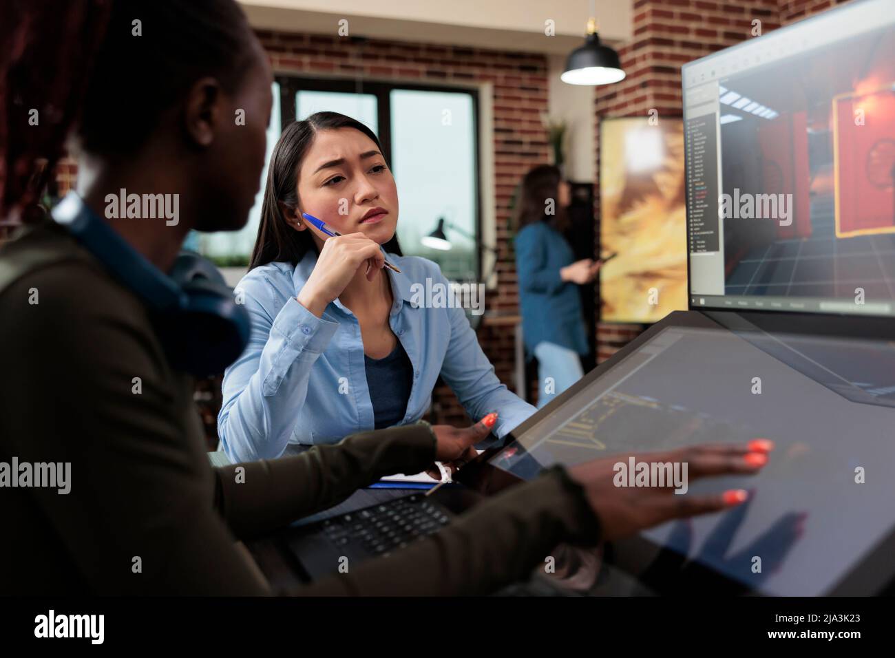Multiethnic creative game creators sitting at desk with multiple displays while working with 3D assets. Digital interactive entertainment industry workers working with CGI. Stock Photo