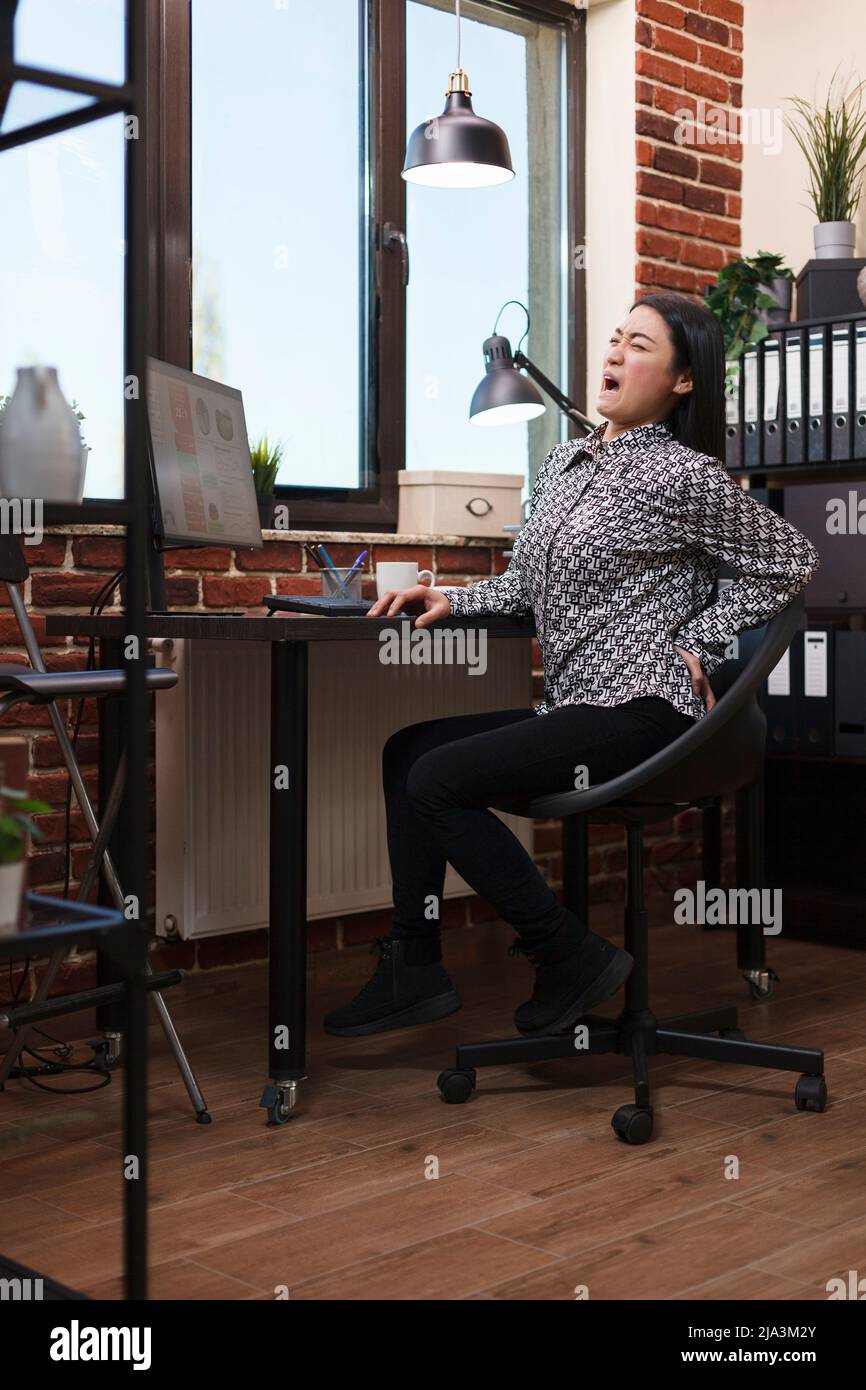 Financial company exhausted businesswoman with serious backache sitting on chair at workplace. Marketing agency office employee accusing painful back sitting at desk in workspace. Stock Photo