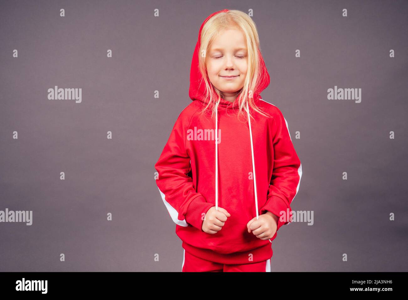 blonde blue-eyed little model girl in a red sports suit posing in the studio on a black background.active childhood full length portrait put on hood Stock Photo