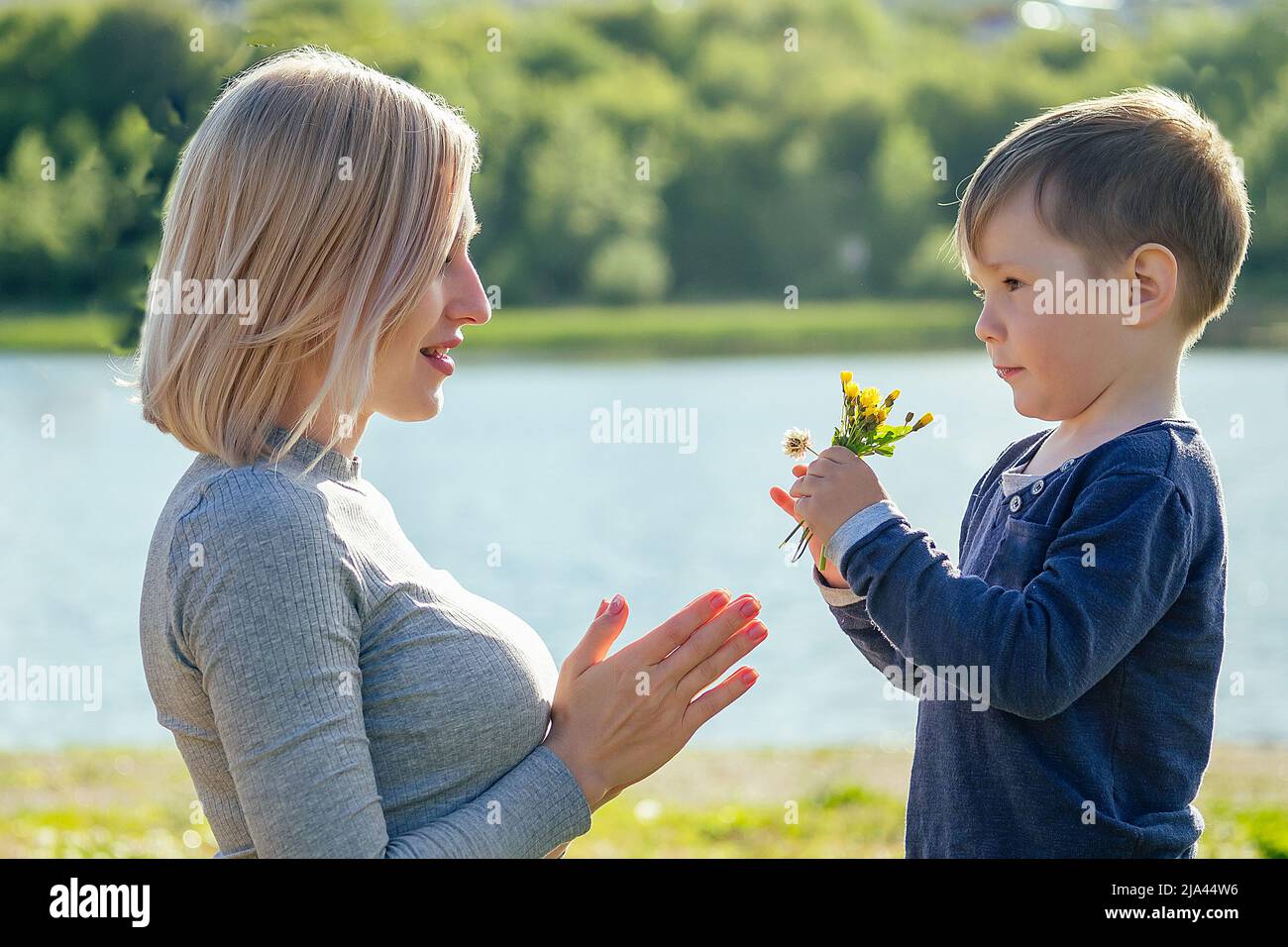 cute baby (son) gives a gift flowers to his beautiful blonde mother in the park on a background of green grass and lake . mother's day concept Stock Photo