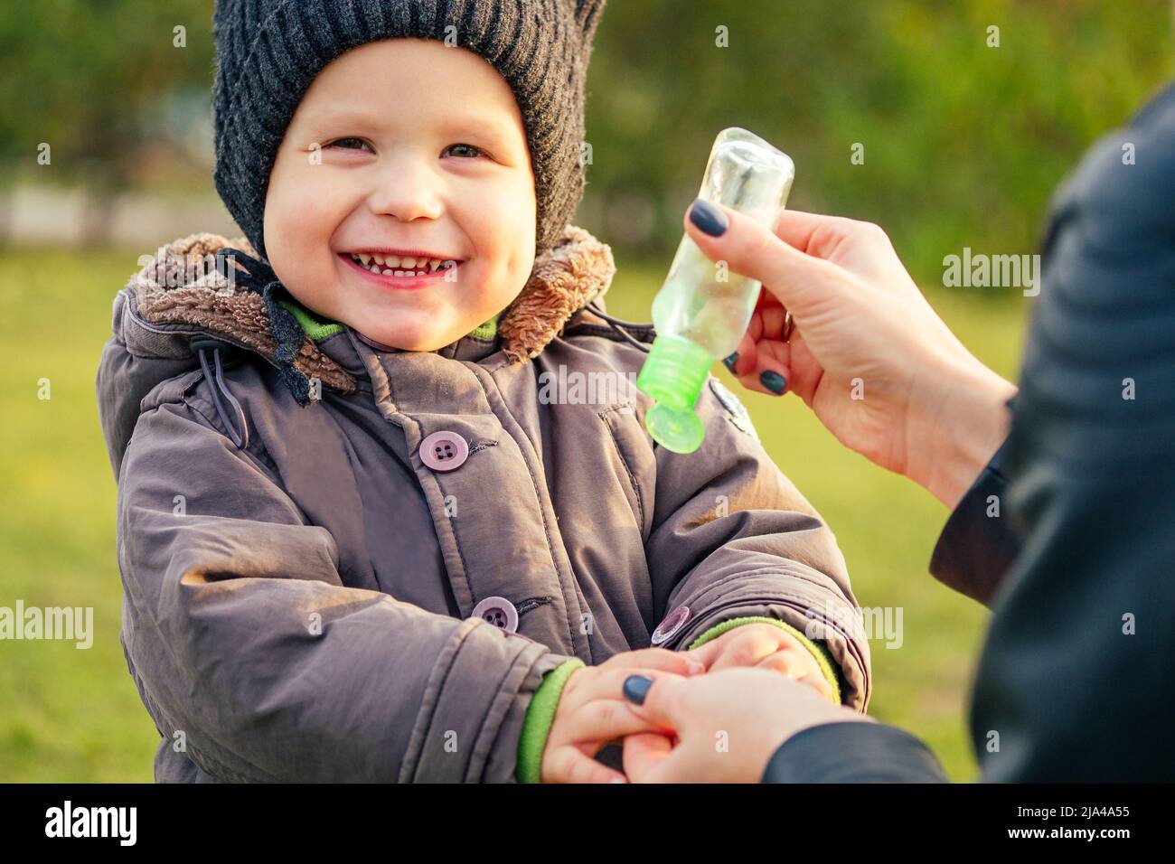 Mother in a warm jacket and knitted hat apply an antiseptic gel ( antibacterial gel ) baby boy walking in the autumn park. stylish blonde woman with a Stock Photo