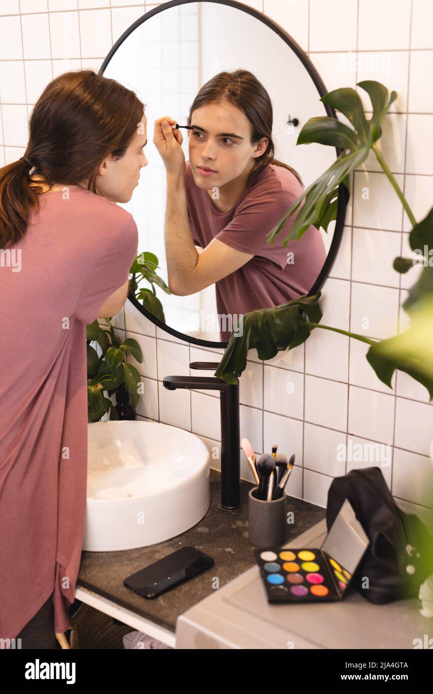 Non-binary trans woman doing her eyebrow makeup looking in the mirror in bathroom at home Stock Photo