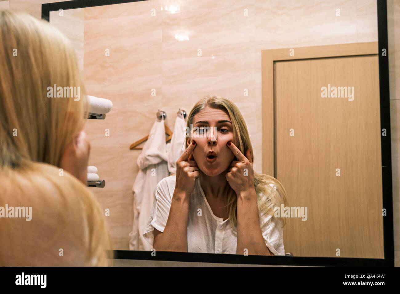 Second chin, obesity. Portrait of 30 year old girl lifting her cheeks up using her fingers while looking at mirror in bathroom. Aging and appearance Stock Photo