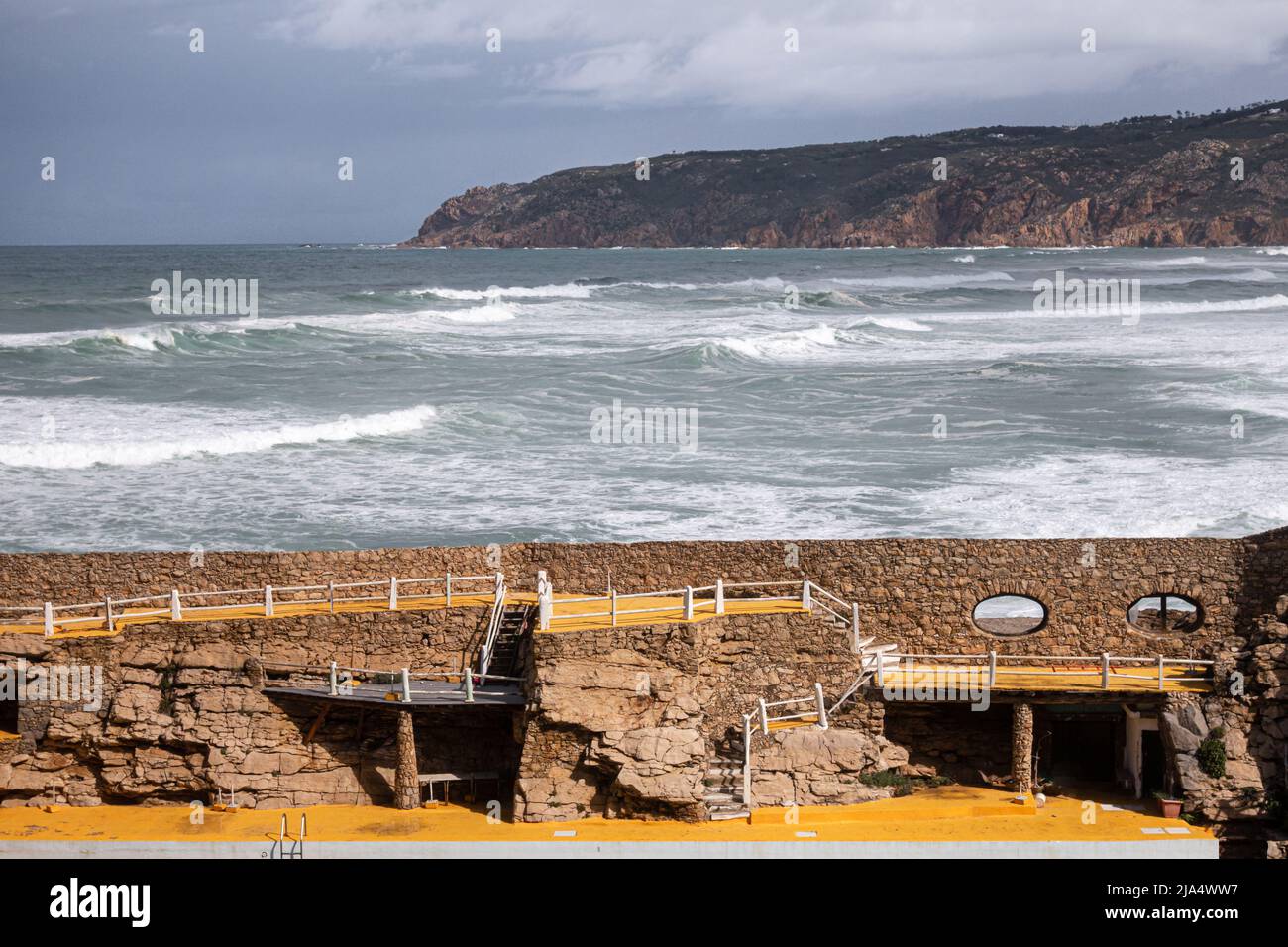 Large Atlantic Ocean waves rolling in to Guincho beach next to tourist pool facility with Cabo Raso and cloud covered horizon background in Cascais Po Stock Photo