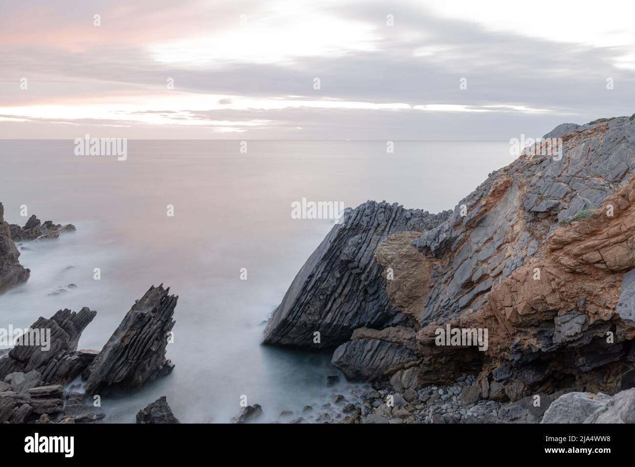 Layered eroded rocks emerging from the Atlantic Ocean at twilight with setting sun at horizon in long exposure landscape in Sintra Portugal Stock Photo