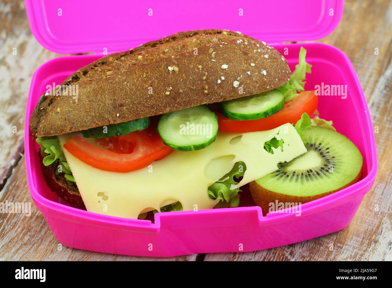 Whole grain roll with cheese, lettuce, tomato and cucumber and kiwi fruit in pink lunch box, closeup Stock Photo