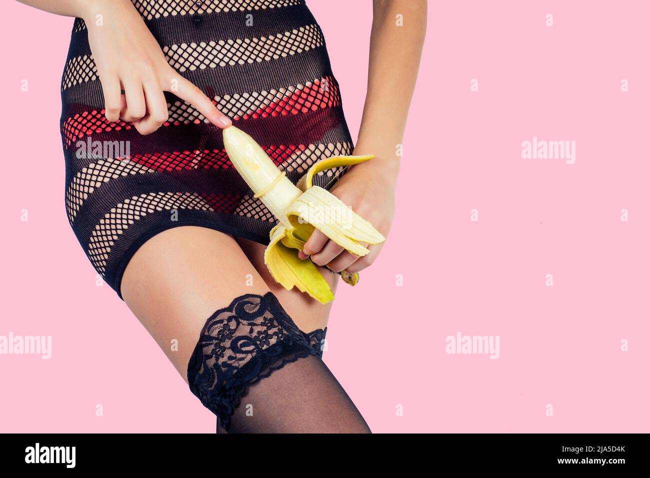 Closeup of a sexy woman body in red panties and black sexy transparent dress stockings touching a banana in a condom on pink background in the studio Stock Photo