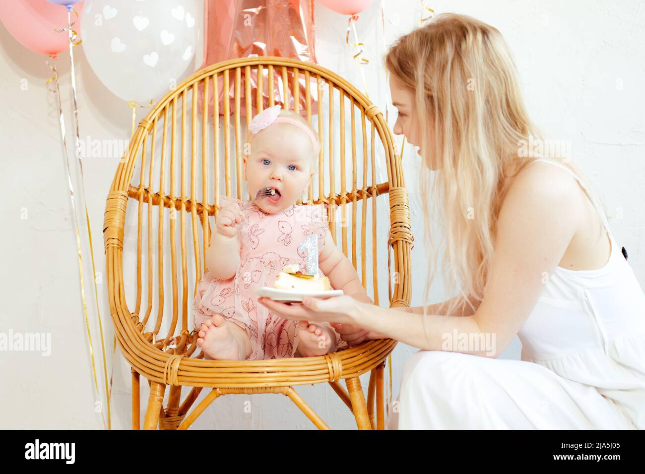Portrait closeup of young mother and baby girl sitting and eating cake in holiday dress on white background. First birthday party, pink decor and Stock Photo