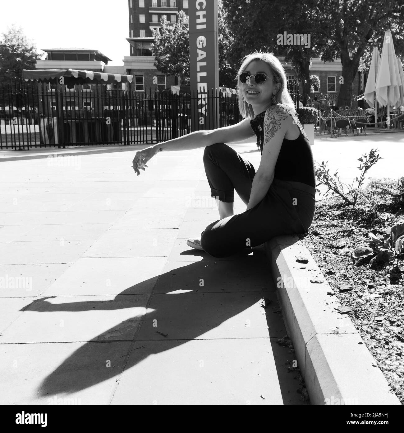 Pretty lady with tattoos, nose piercing and sunglasses sits on a curb stone, smiles and smokes. Duke of York Square, Chelsea, London. Monochrome. Stock Photo