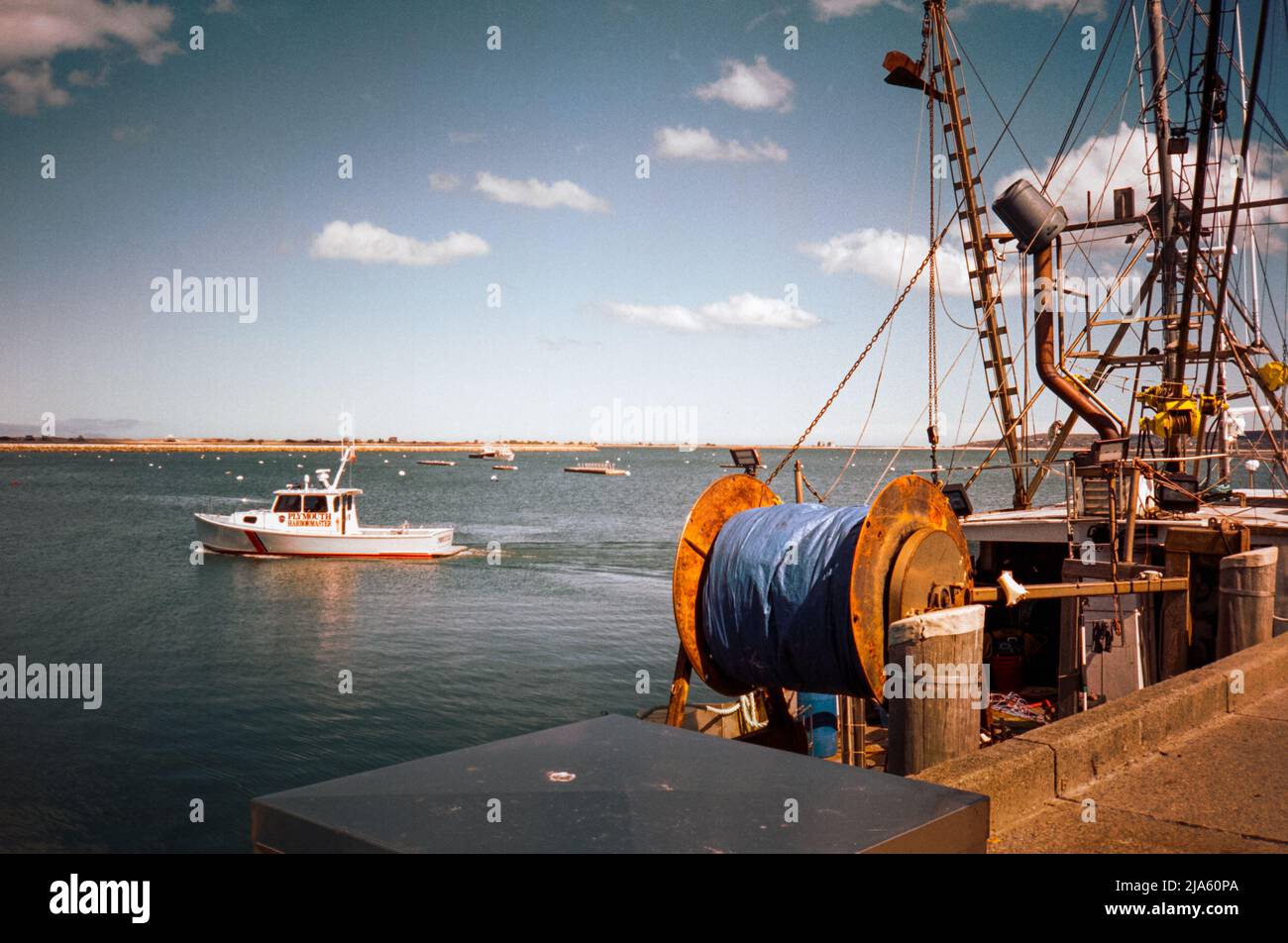 A fishing boat waits at the dock as the Harbor Master boat passes by in the background at Plymouth Massachusetts. The image was captured on analog col Stock Photo