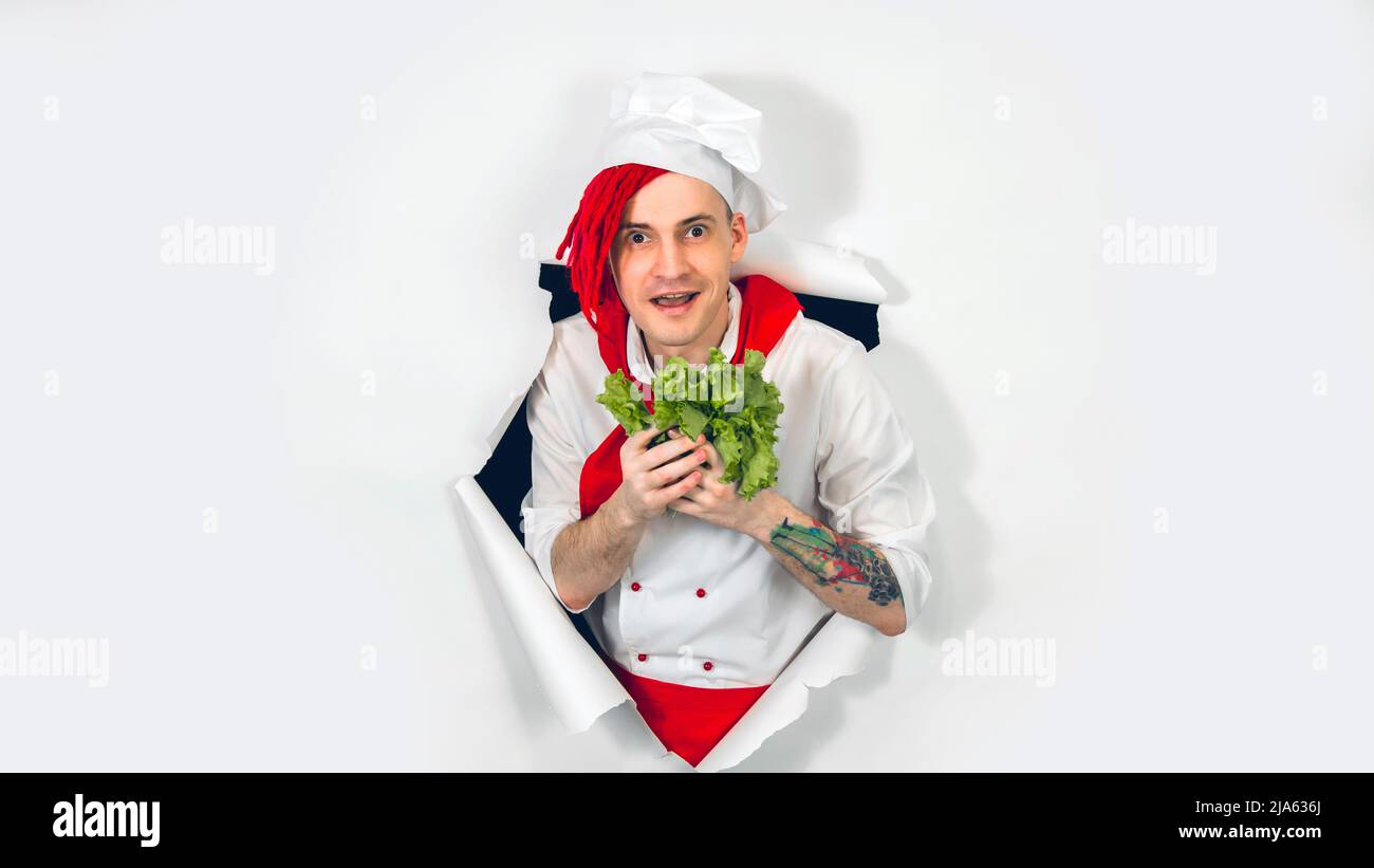 Man with red dreadlocks holds it in his hand lettuce leaves. Cook in white apron and red tie holds an ingredient for a vegetarian dish in his hand, pe Stock Photo