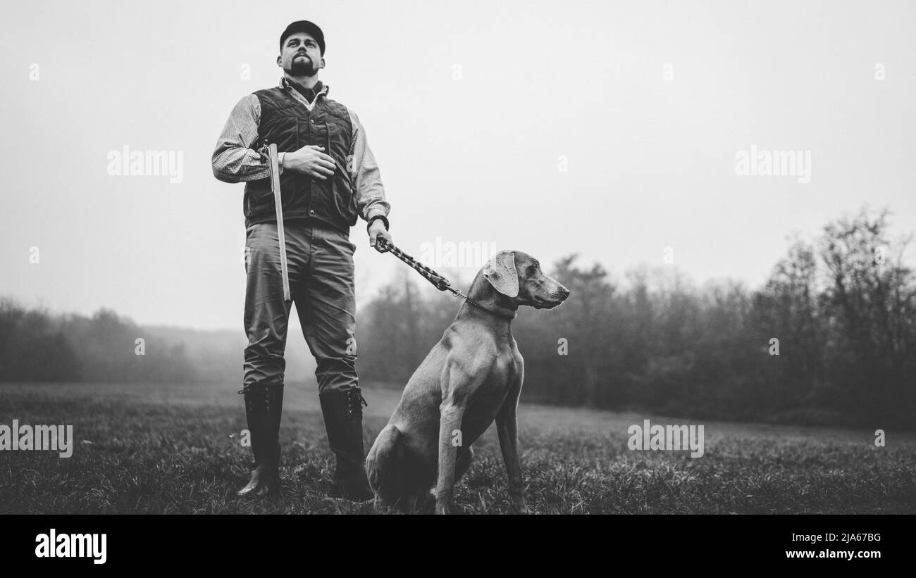 Hunter man with dog in traditional shooting clothes on field holding shotgun, black and white photo. Stock Photo