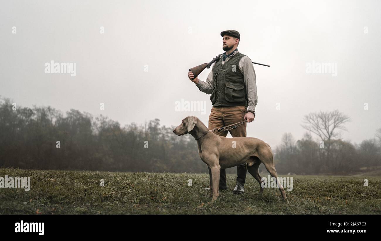 Hunter man with dog in traditional shooting clothes on field holding shotgun. Stock Photo