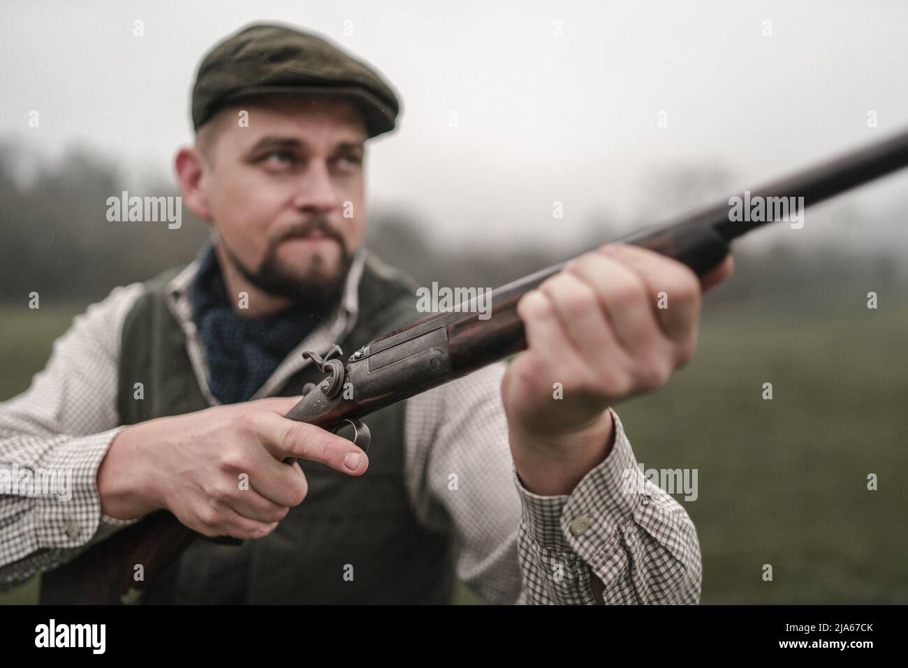 Hunter man in traditional shooting clothes on field aiming with shotgun