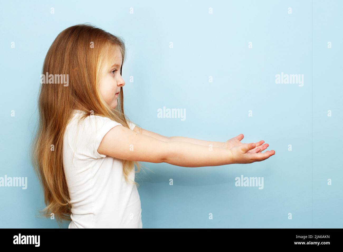 little blonde girl with long hair stretched her arms to the side. child holding open palm empty hand on a blue background Stock Photo