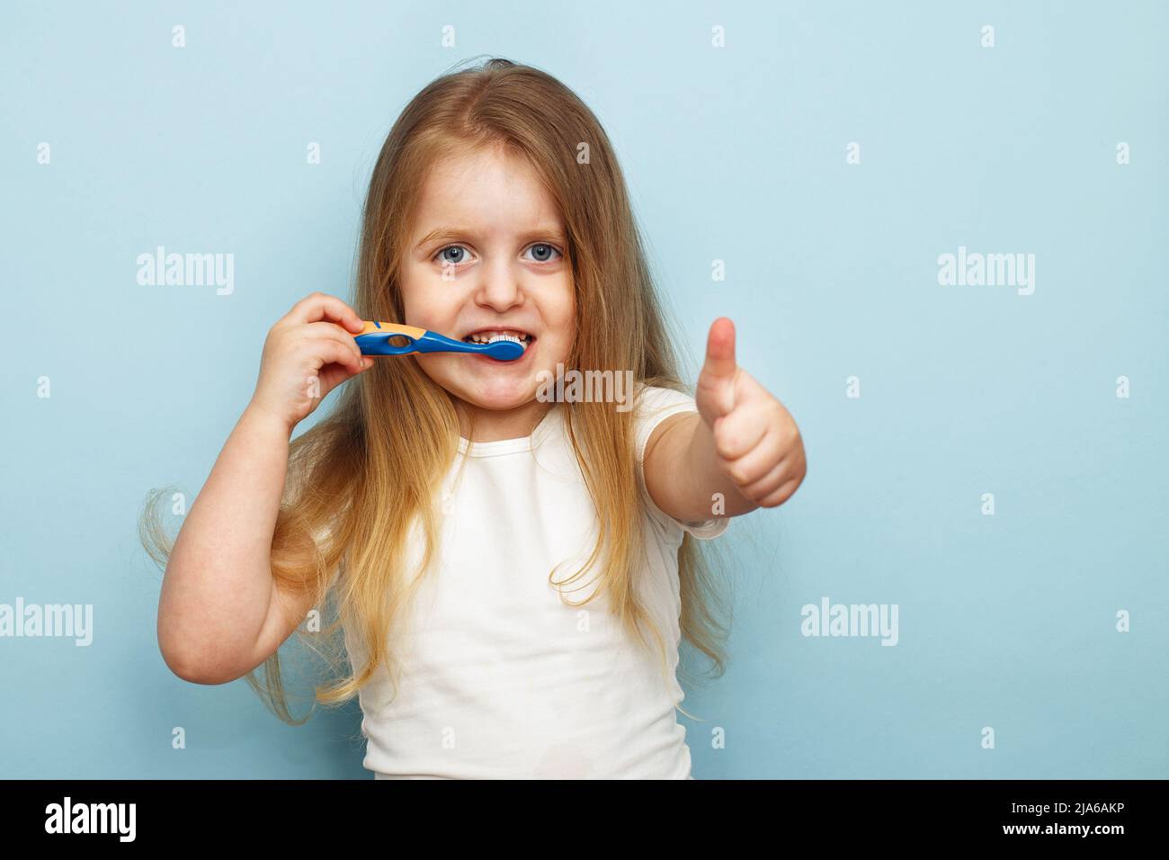little happy girl brushing her teeth with a toothbrush and smiling on a blue background. thumb up Stock Photo
