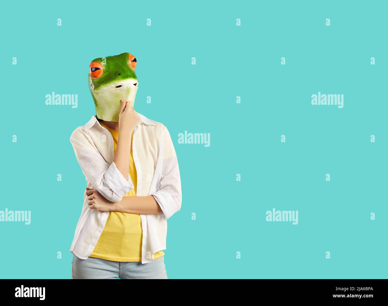 Funny woman in rubber mask of frog stands in pensive pose on turquoise background. Stock Photo